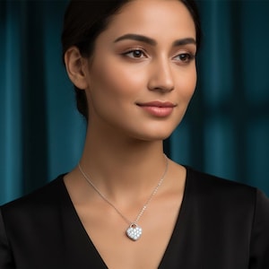 Könnte beinhalten: Eine silberne Halskette mit einem herzförmigen Anhänger. Der Anhänger ist mit kleinen, klaren Edelsteinen besetzt. Die Frau im Bild trägt ein schwarzes Oberteil.