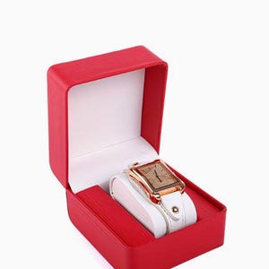 May include: A rectangular wristwatch with a rose gold-tone case and a white leather strap, displayed inside an open, bright red presentation box with a white interior lining. The watch face features a simple dial with thin, dark hour markers.