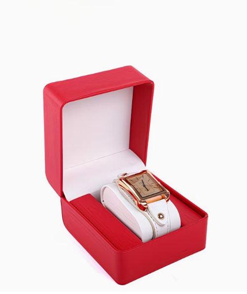 May include: A rectangular wristwatch with a rose gold-tone case and a white leather strap, displayed inside an open, bright red presentation box with a white interior lining. The watch face features a simple dial with thin, dark hour markers.