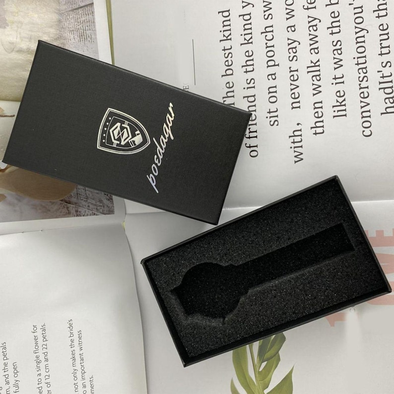 May include: A rectangular black watch box with a lid featuring a white shield logo and the text poedagar. The open base contains a black foam insert with a cutout shaped like a wristwatch, designed to hold a timepiece securely within the packaging.