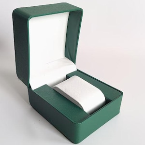 May include: An open rectangular watch box featuring a textured dark green exterior and a smooth white interior lining. The box includes a central white padded watch cushion designed to hold a timepiece securely within the structured green casing.