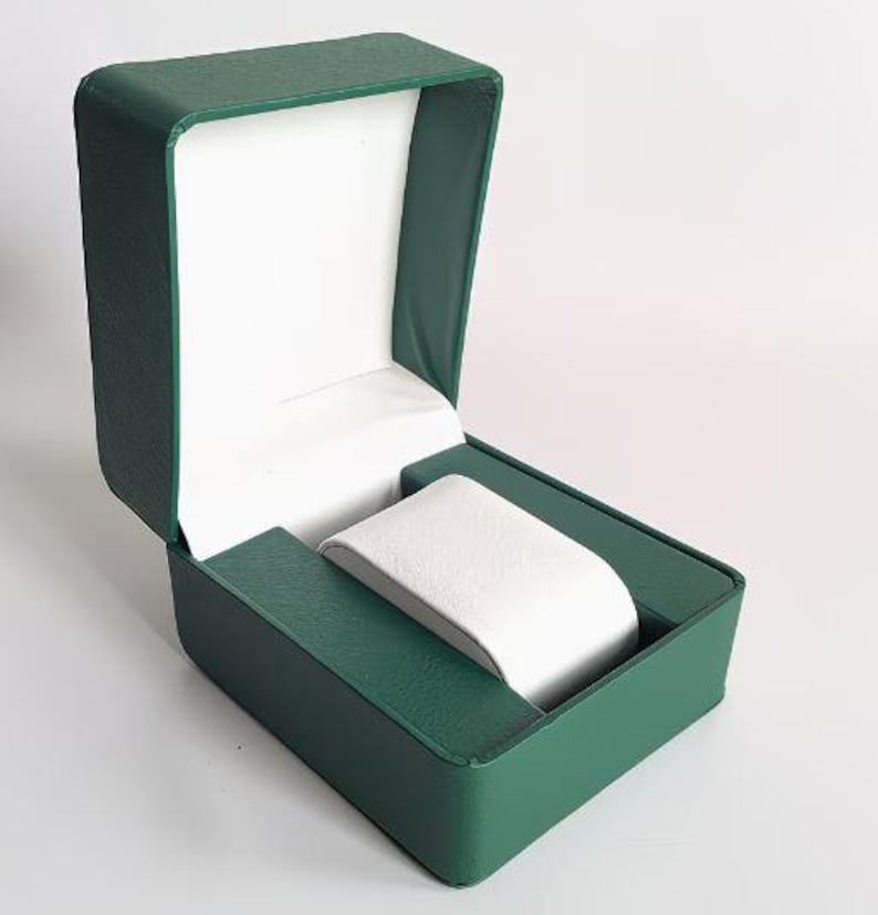 May include: An open rectangular watch box featuring a textured dark green exterior and a smooth white interior lining. The box includes a central white padded watch cushion designed to hold a timepiece securely within the structured green casing.