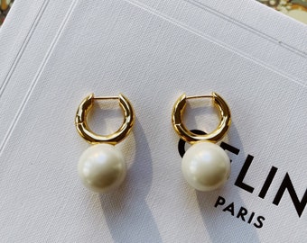 Puede incluir: Un par de pendientes de aro dorados con una perla colgante. Los pendientes están sobre una superficie blanca con el texto "CELINE PARIS" visible.