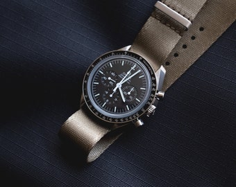 Könnte beinhalten: Eine schwarze Armbanduhr mit silbernem Gehäuse und einem khakifarbenen Nylonarmband. Das Zifferblatt der Uhr hat mehrere Hilfszifferblätter und weiße Zeiger. Die Uhr wird auf einer dunkelblauen, strukturierten Oberfläche präsentiert.