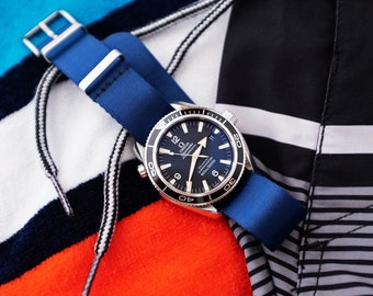 May include: A luxury wristwatch with a blue face and a blue nylon strap. The watch has a black bezel with white markings and silver hands. The watch is resting on a striped fabric.