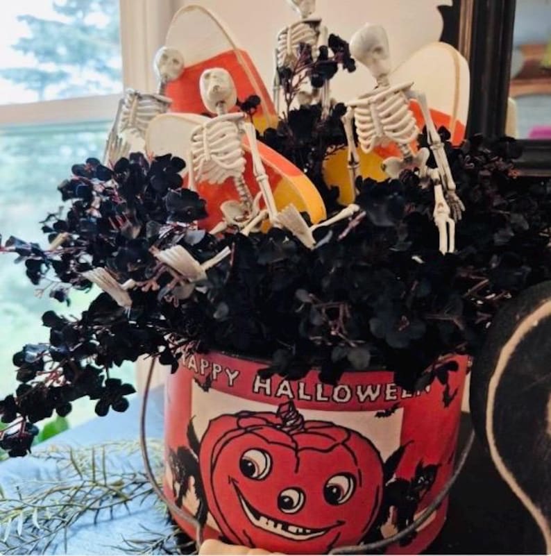 Puede incluir: Un cubo tem&aacute;tico de Halloween con un dise&ntilde;o de calabaza sonriente y las palabras "Happy Halloween". El cubo est&aacute; lleno de follaje negro, recortes de caramelos y peque&ntilde;as figuras de esqueletos, creando una exhibici&oacute;n decorativa espeluznante.