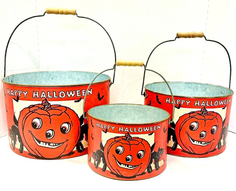 Puede incluir: Conjunto de tres cubos de Halloween de estilo vintage. Cada cubo rojo presenta un dise&ntilde;o de calabaza sonriente y las palabras "Happy Halloween". Tienen asas de alambre negro con empu&ntilde;aduras de madera, ideales para caramelos.