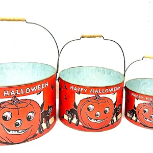 Puede incluir: Juego de tres cubos de Halloween con asas de alambre y empu&ntilde;aduras de madera. Los cubos rojos presentan una calabaza sonriente, gatos negros y las palabras "Happy Halloween". Los cubos son de diferentes tama&ntilde;os.