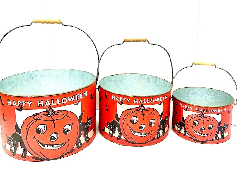 Puede incluir: Juego de tres cubos de Halloween con asas de alambre y empu&ntilde;aduras de madera. Los cubos rojos presentan una calabaza sonriente, gatos negros y las palabras "Happy Halloween". Los cubos son de diferentes tama&ntilde;os.