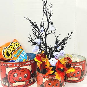 Puede incluir: Cubos de metal con tem&aacute;tica de Halloween con un dise&ntilde;o de calabaza sonriente. Un cubo contiene una bolsa de Cheetos Puffs. Un &aacute;rbol negro con luces de fantasma y decoraci&oacute;n de hojas de oto&ntilde;o decora la escena. Los cubos son rojos con la inscripci&oacute;n "Happy Halloween".