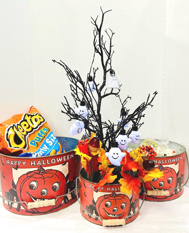 Puede incluir: Cubos de metal con tem&aacute;tica de Halloween con un dise&ntilde;o de calabaza sonriente. Un cubo contiene una bolsa de Cheetos Puffs. Un &aacute;rbol negro con luces de fantasma y decoraci&oacute;n de hojas de oto&ntilde;o decora la escena. Los cubos son rojos con la inscripci&oacute;n "Happy Halloween".