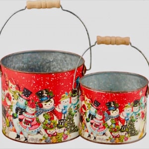 May include: Two red metal buckets with snowman designs and wooden handles. The buckets are nested, with the smaller bucket inside the larger bucket.