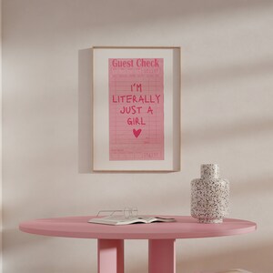 K&ouml;nnte beinhalten: Ein rosa gerahmter Druck mit dem Text "Guest Check" und "I'm Literally Just A Girl" mit einem rosa Herzen. Der Druck befindet sich auf einem rosa Tisch mit einer wei&szlig;-schwarz gefleckten Vase.