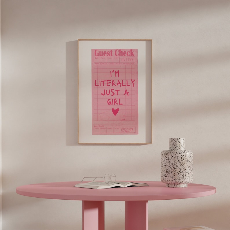 K&ouml;nnte beinhalten: Ein rosa gerahmter Druck mit dem Text "Guest Check" und "I'm Literally Just A Girl" mit einem rosa Herzen. Der Druck befindet sich auf einem rosa Tisch mit einer wei&szlig;-schwarz gefleckten Vase.