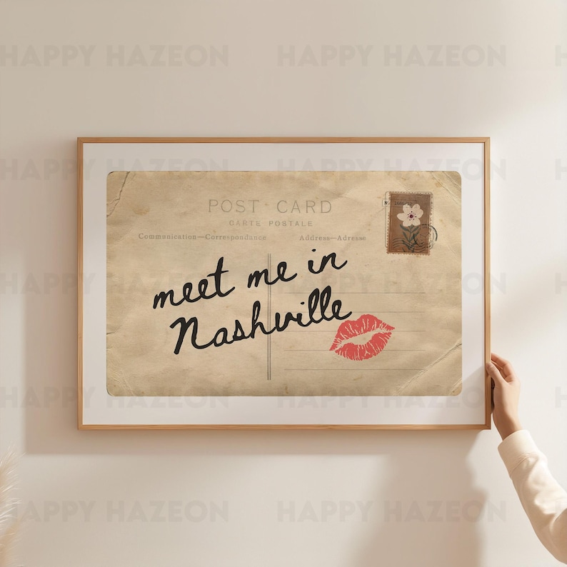 May include: A framed vintage postcard print with the text "meet me in Nashville" in black script, a red lip print, and a postage stamp. The postcard has a sepia tone and is set in a wooden frame.
