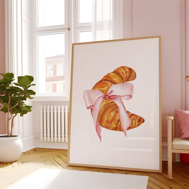 May include: A watercolor painting of a croissant tied with a pink ribbon. The croissant is a light brown color and the ribbon is a pale pink. The painting is on a white background.