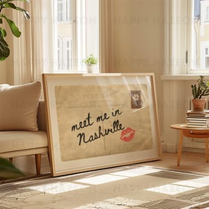 May include: A framed vintage postcard print with the text "meet me in Nashville" in black script and a red lipstick kiss. The print is in a light wood frame, leaning against a wall.