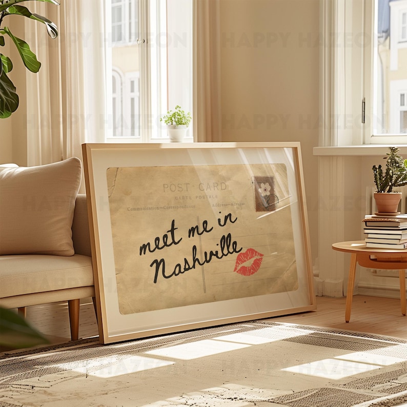 May include: A framed vintage postcard print with the text "meet me in Nashville" in black script and a red lipstick kiss. The print is in a light wood frame, leaning against a wall.