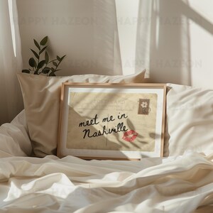 May include: A framed vintage postcard with the text "meet me in Nashville" in cursive script, a red lip print, and a floral stamp. The postcard is set against a backdrop of soft, cream-colored bedding.