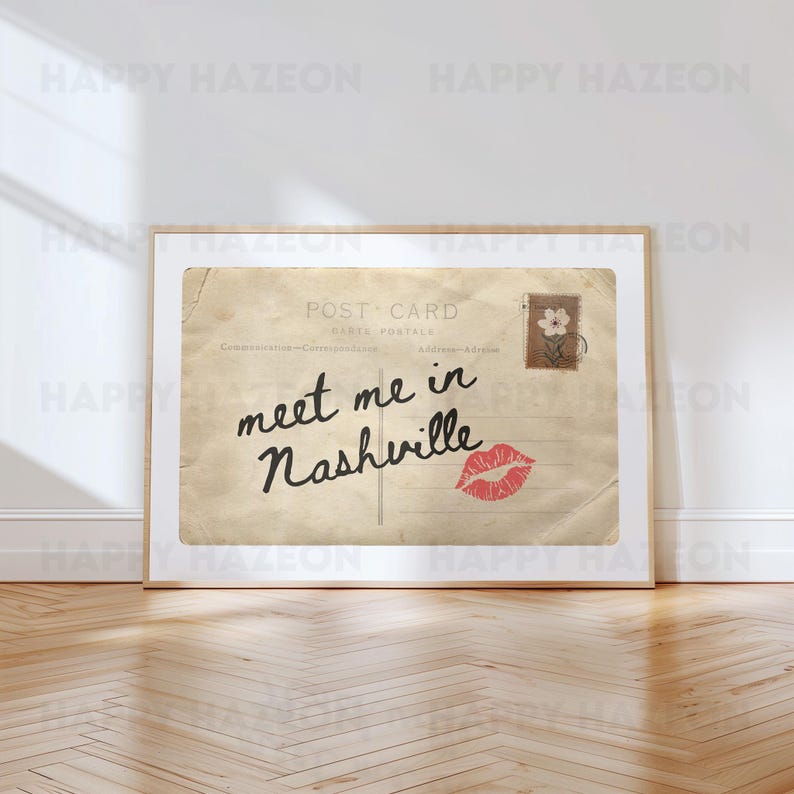 May include: A framed vintage postcard with the text "meet me in Nashville" in cursive. The postcard has a red kiss mark and a postage stamp with a flower. The frame is a light brown color.