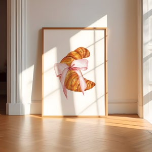 May include: A watercolor illustration of a croissant tied with a pink ribbon. The croissant is a light brown color and the ribbon is a pale pink. The illustration is on a white background.