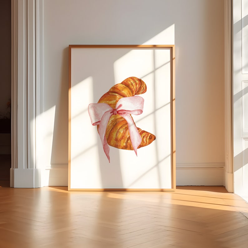 May include: A watercolor illustration of a croissant tied with a pink ribbon. The croissant is a light brown color and the ribbon is a pale pink. The illustration is on a white background.