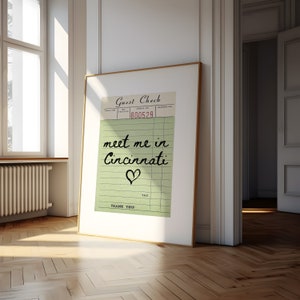 May include: A framed print with a green and white guest check design. The text "meet me in Cincinnati" is written in black cursive with a heart symbol below. The check number is 800529.