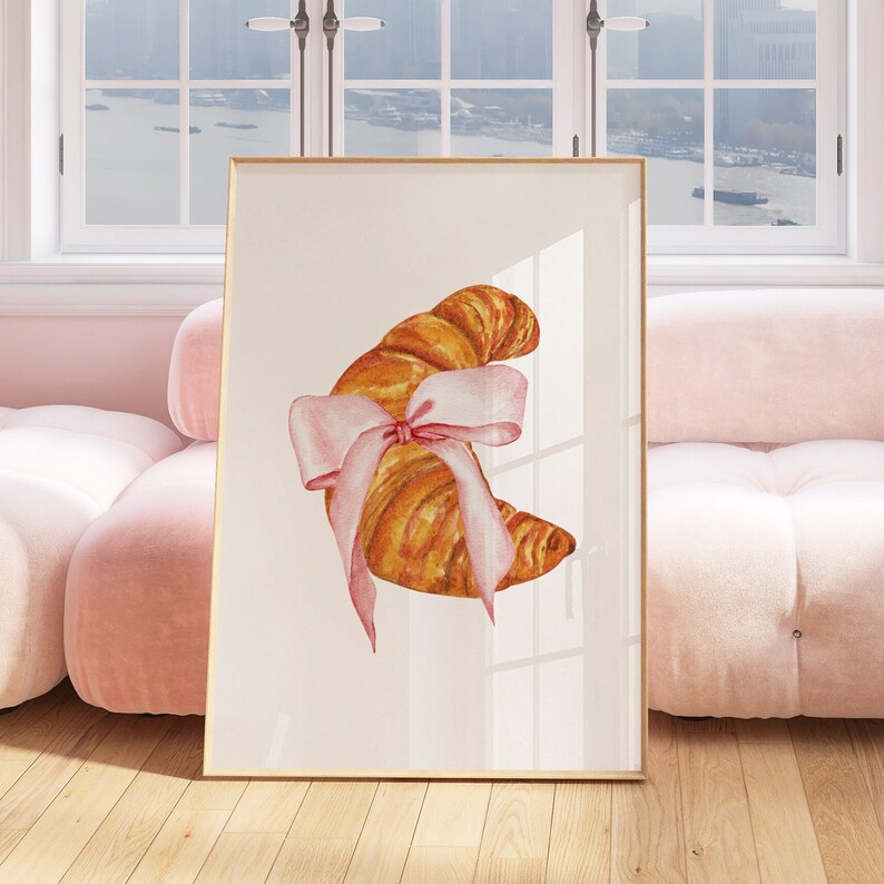 May include: A watercolor illustration of a croissant tied with a pink ribbon. The croissant is a crescent shape and is a light brown color. The ribbon is tied in a bow and is a light pink color.