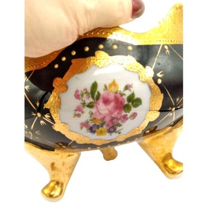 May include: A black and gold porcelain bowl with a floral design. The bowl has three gold feet and a white center with a pink rose and other flowers.