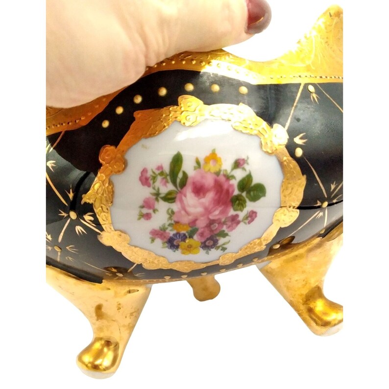 May include: A black and gold porcelain bowl with a floral design. The bowl has three gold feet and a white center with a pink rose and other flowers.