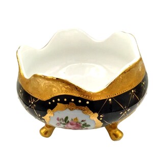 May include: A small, footed bowl with a black and gold design. The bowl has a white interior and a floral design on the front. The bowl is made of porcelain.
