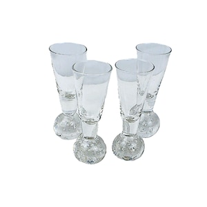 Op de afbeelding: Vier heldere shotglaasjes met een uniek ontwerp. Elk glas heeft een ronde, bubbelachtige voet en een uitlopende bovenkant. De glazen staan op een witte achtergrond.