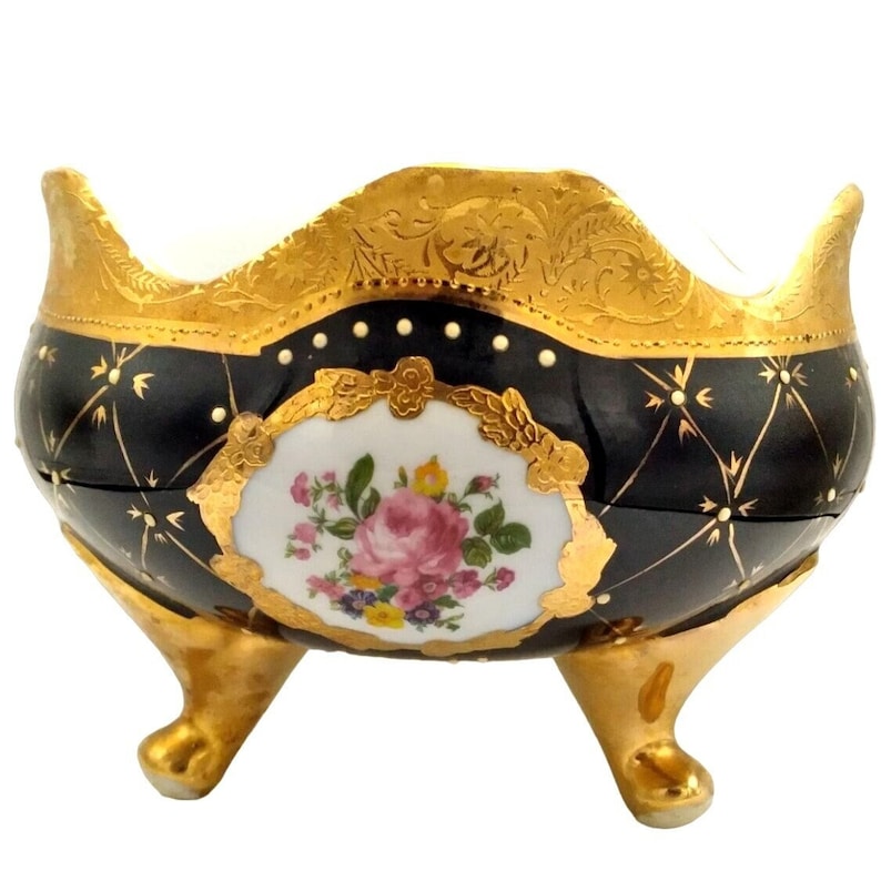 May include: A black and gold porcelain bowl with a floral design and three legs. The bowl has a scalloped rim and is decorated with gold accents.