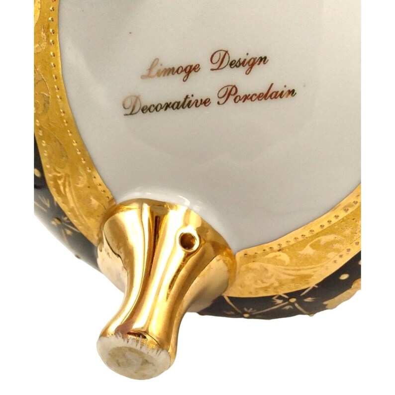 May include: Close-up of the bottom of a black and gold decorative porcelain egg. The egg has a gold trim and a gold foot. The inscription on the bottom reads "Limoge Design Decorative Porcelain".
