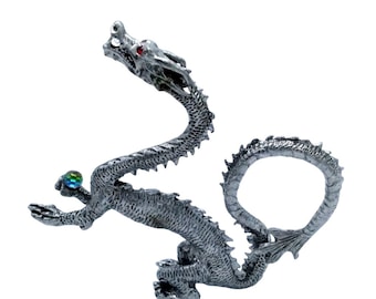 May include: A silver-colored dragon figurine with a long, serpentine body and a curved tail. The dragon holds a small, iridescent orb in its clawed hand. The dragon has red eyes and is set against a white background.