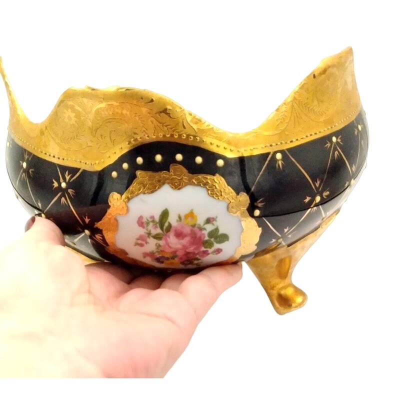 May include: A black and gold porcelain bowl with a floral design and three feet. The bowl has a scalloped edge and is decorated with gold accents.