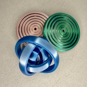 May include: Three 3D-printed objects in shades of blue, green, and pink. The blue object is a Mobius strip design. The green and pink objects are circular with concentric rings. The objects are on a light gray surface.