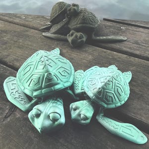 May include: Three sea turtle figurines with textured, geometric shell patterns. Two turtles are light mint green, while one turtle in the background is a dark, muted olive green. Each turtle features a detailed head and flippers.