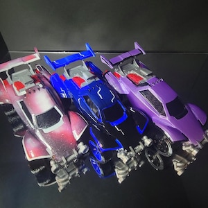 May include: Three toy race cars with rear spoilers and detailed chassis designs. The left car features a pink and white nebula pattern, the center car is blue with black accents, and the right car is solid purple. Text reads: This listing is for 1 car.