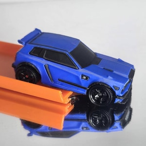 Può includere: Un'auto giocattolo blu con finestrini e ruote nere, posizionata su una pista di plastica arancione. Il veicolo presenta una carrozzeria di tipo hatchback con uno spoiler posteriore e finiture nere lungo i lati e il cofano.