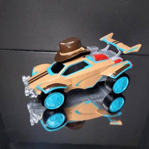 Może przedstawiać: Beżowy samochodzik z niebieskimi i brązowymi akcentami, tylnym spojlerem i brązowym kapeluszem kowbojskim na górze. Koła są czarne z niebieskimi środkami. Samochód odbija się na błyszczącej czarnej powierzchni.