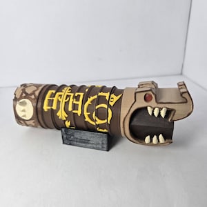 Peut inclure: Un objet cylindrique marron orné de symboles jaunes en relief et d'une tête de créature sculptée à une extrémité avec des dents blanches et un œil rouge. L'objet repose sur un petit support rectangulaire noir et présente une texture.