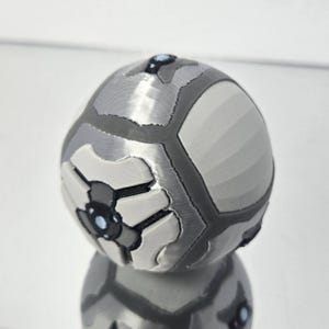May include: A grey and white spherical object with a geometric design. The object has a segmented appearance with dark grey outlines and white and black accents. The top has a small, dark grey detail. The object appears to be 3D-printed.