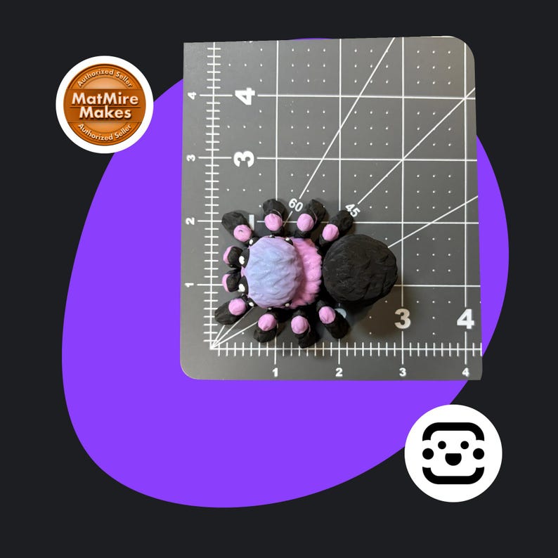 May include: A handmade spider figurine with a black body and legs, and a light blue and pink abdomen. The spider is approximately 4 cm in length and is placed on a gray cutting mat. The image includes the text "MatMire Makes".