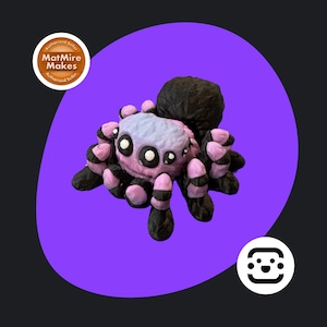 May include: A handmade spider ornament with a black body and legs, and a pink and lilac head. The spider has white eyes and is set against a purple backdrop. The figurine is a unique decorative item.