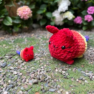 Puede incluir: Dos juguetes de gato hechos a mano con ganchillo. El gato más grande es rojo con detalles de arcoíris y una pequeña cola azul. El gato más pequeño es rojo con detalles de arcoíris y una cola azul. Los juguetes están sobre un lecho de pequeñas rocas y hierba verde.