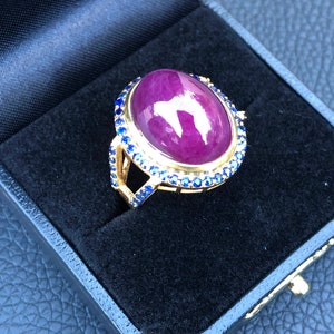 Puede incluir: Un anillo de oro con una gran piedra de rubí ovalada rodeada por un halo de pequeños zafiros azules.