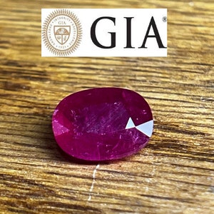 May include: A close-up of a deep red oval-shaped ruby gemstone. The ruby is on a wooden surface. The gemstone is certified by the Gemological Institute of America (GIA).