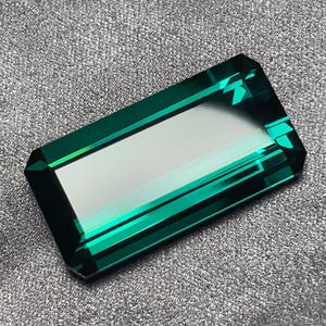 May include: A deep green gemstone with a rectangular shape and a faceted cut. The gemstone is cut with a high level of precision and has a brilliant shine.