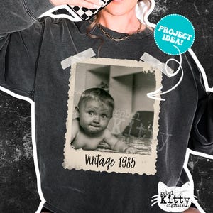 May include: Dark gray sweatshirt featuring a vintage photo print of a baby with the text "Vintage 1985." The photo is taped to the sweatshirt with white tape. A teal circle with the words "PROJECT IDEA!" is also on the sweatshirt.
